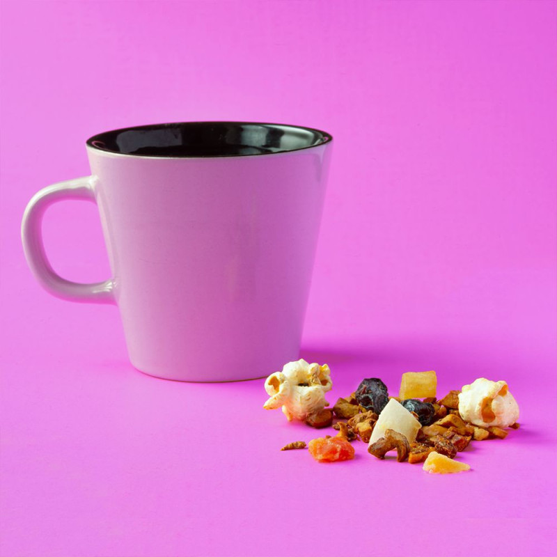 Weiße Tasse mit Kräutertee-Aufguss „Mandelwolke“, getrocknete Früchtestücke und Popcorn auf rosa Hintergrund