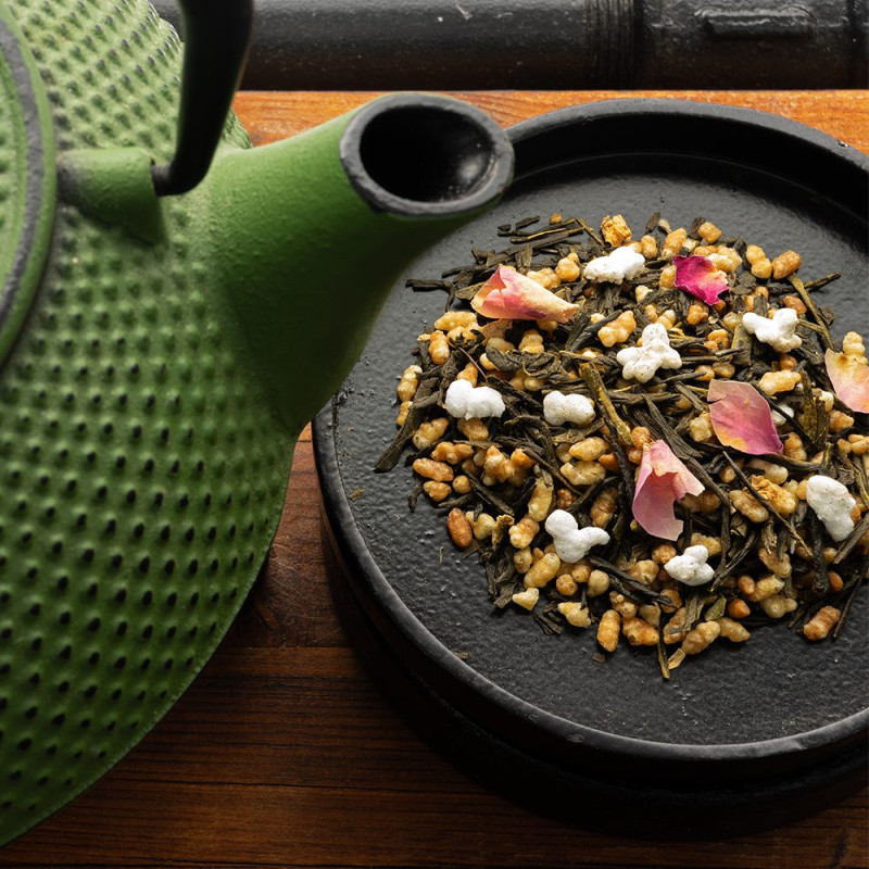 Japanese green tea with rose petals and puffed rice grains on black tray next to green teapot