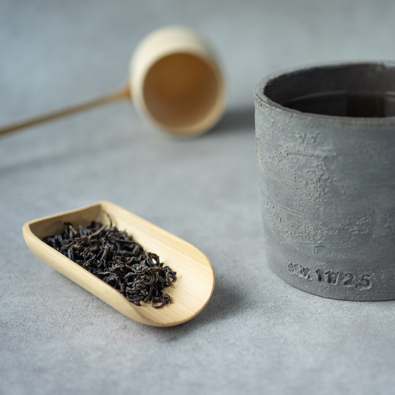 Feuilles de thé noir Jardin d'Alice dans une cuillère en bois à côté d'une tasse en béton