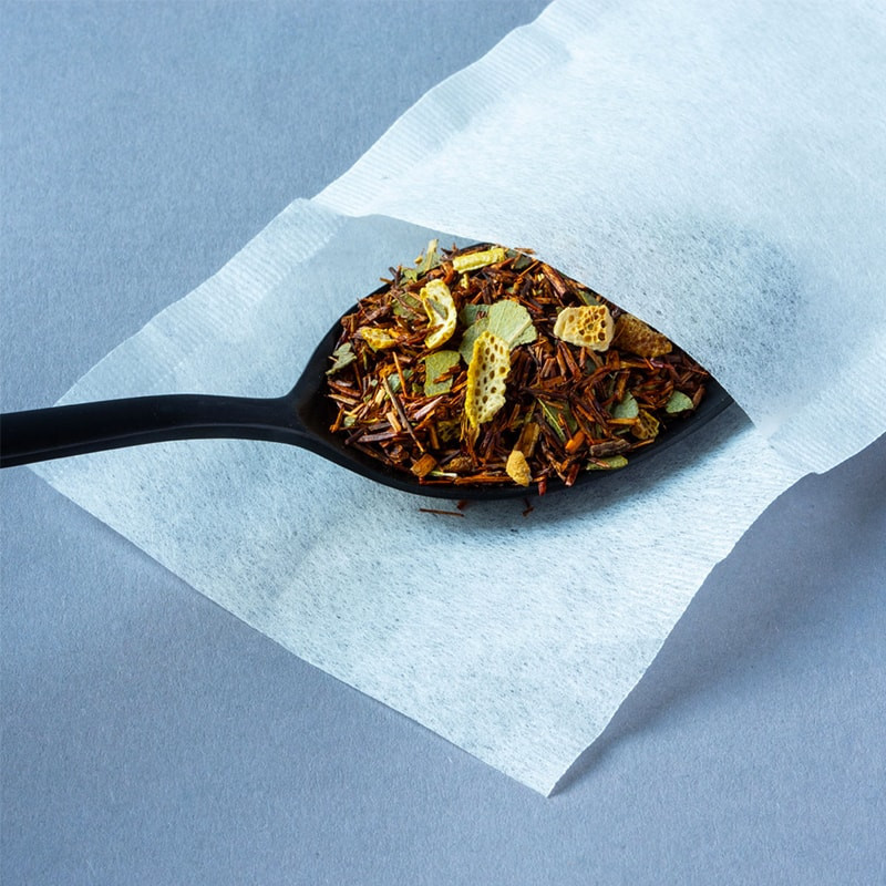 Leeg wit papieren theezakje voor infusie met bladeren en stukjes citrusvruchten op zwarte lepel