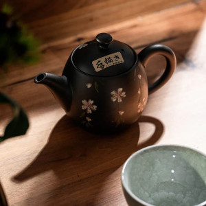 Schwarze Tokoname-Teekanne mit weißen Blütenmustern auf Holztisch, daneben grüne Keramiktasse