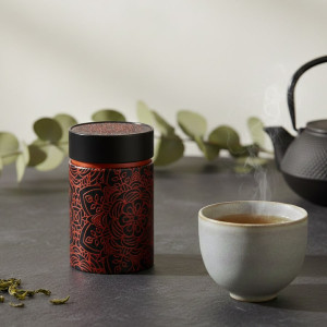 Schwarze Teedose Mandala Ebony mit roten Blütenmotiven auf schwarzem Grund neben einer dampfenden Teetasse