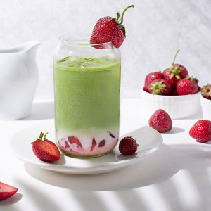 Glas Matcha-Latte mit Erdbeere, Erdbeerstücken und Eiswürfeln auf weißem Teller