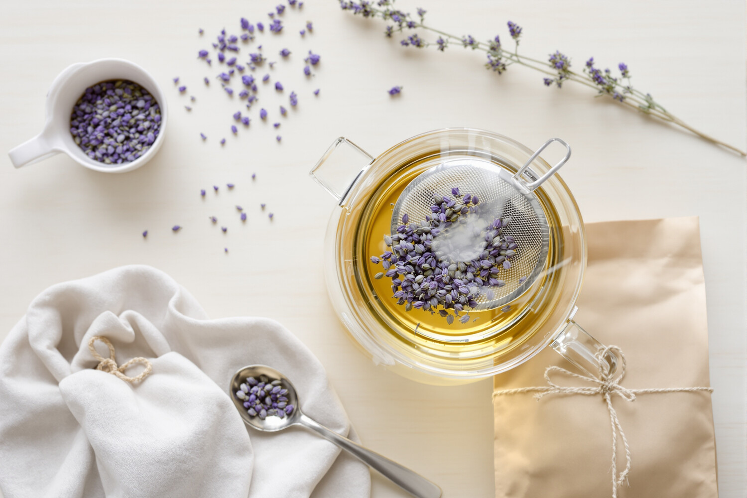 Théière en verre avec fleurs de lavande en infusion et cuillère argentée sur bois clair.